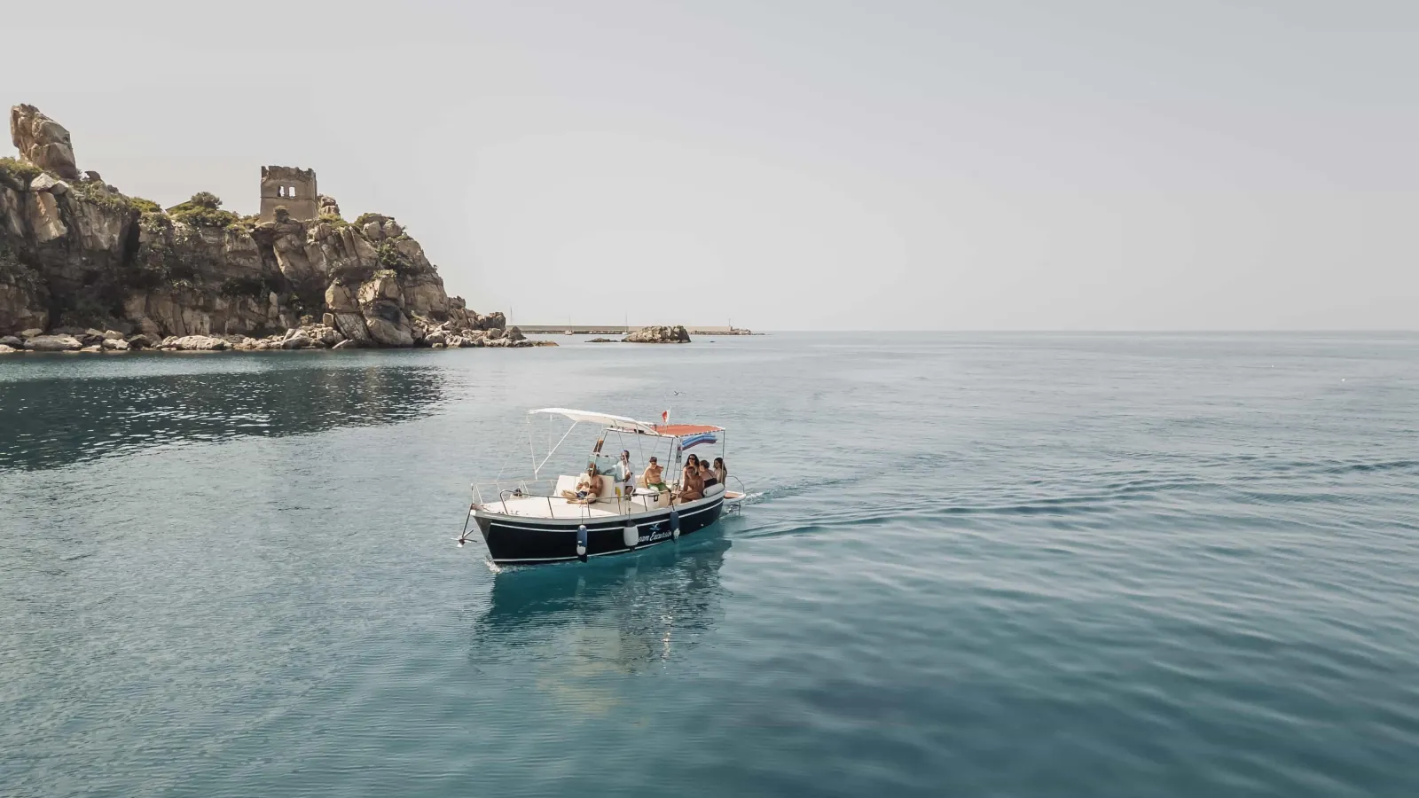 The boat under way along the Cefalù coast with guests aboard, the headland in the haze behind