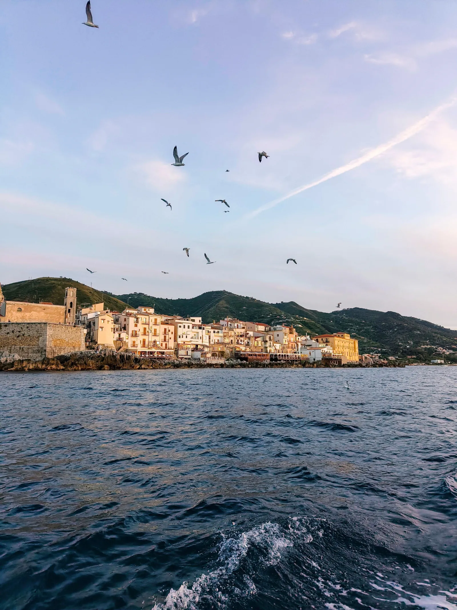 Cefalù's old town in silhouette at dusk, seen from the water with gulls overhead