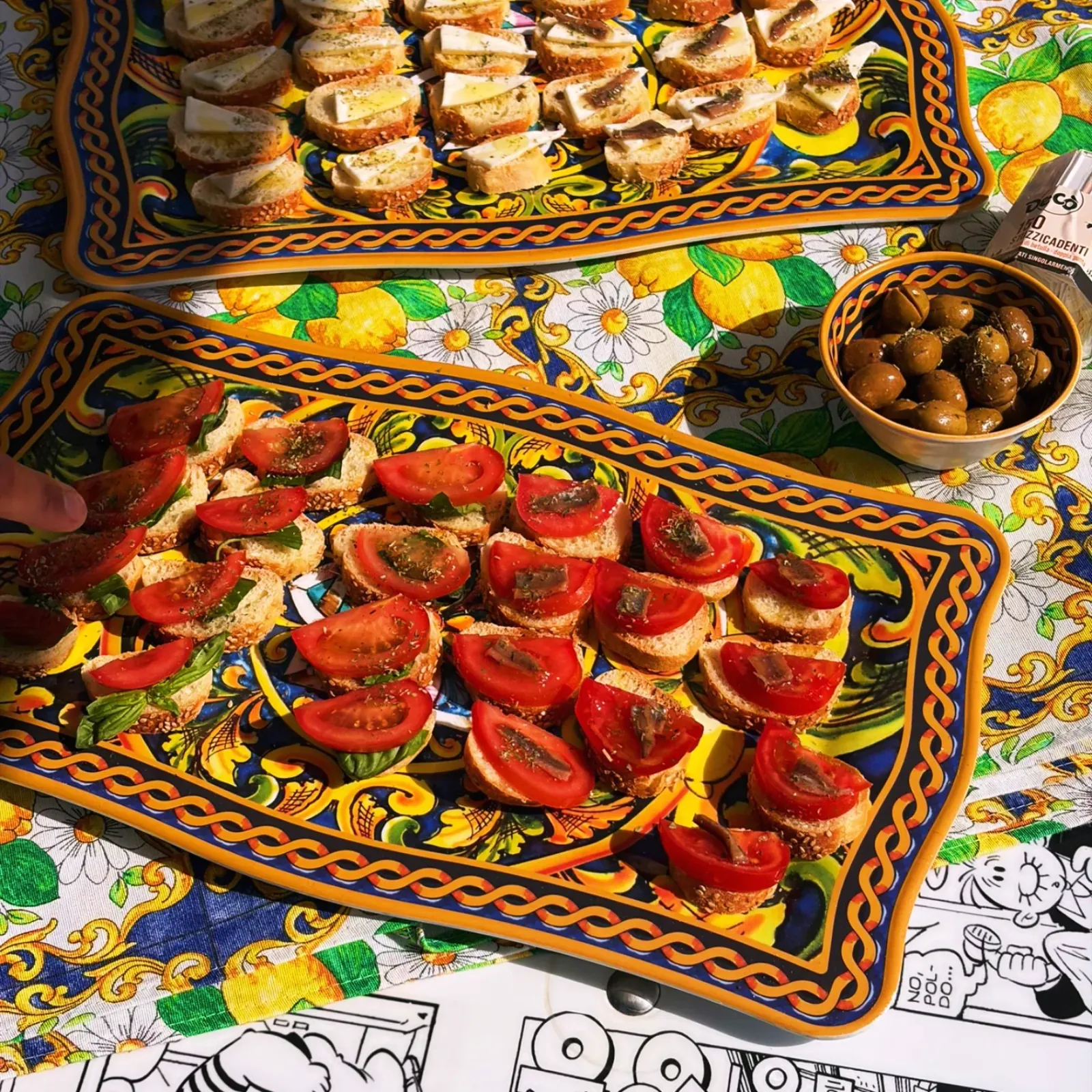 The Sicilian aperitivo laid out on a painted ceramic tray: tomato bruschetta, cured toppings and a bowl of olives