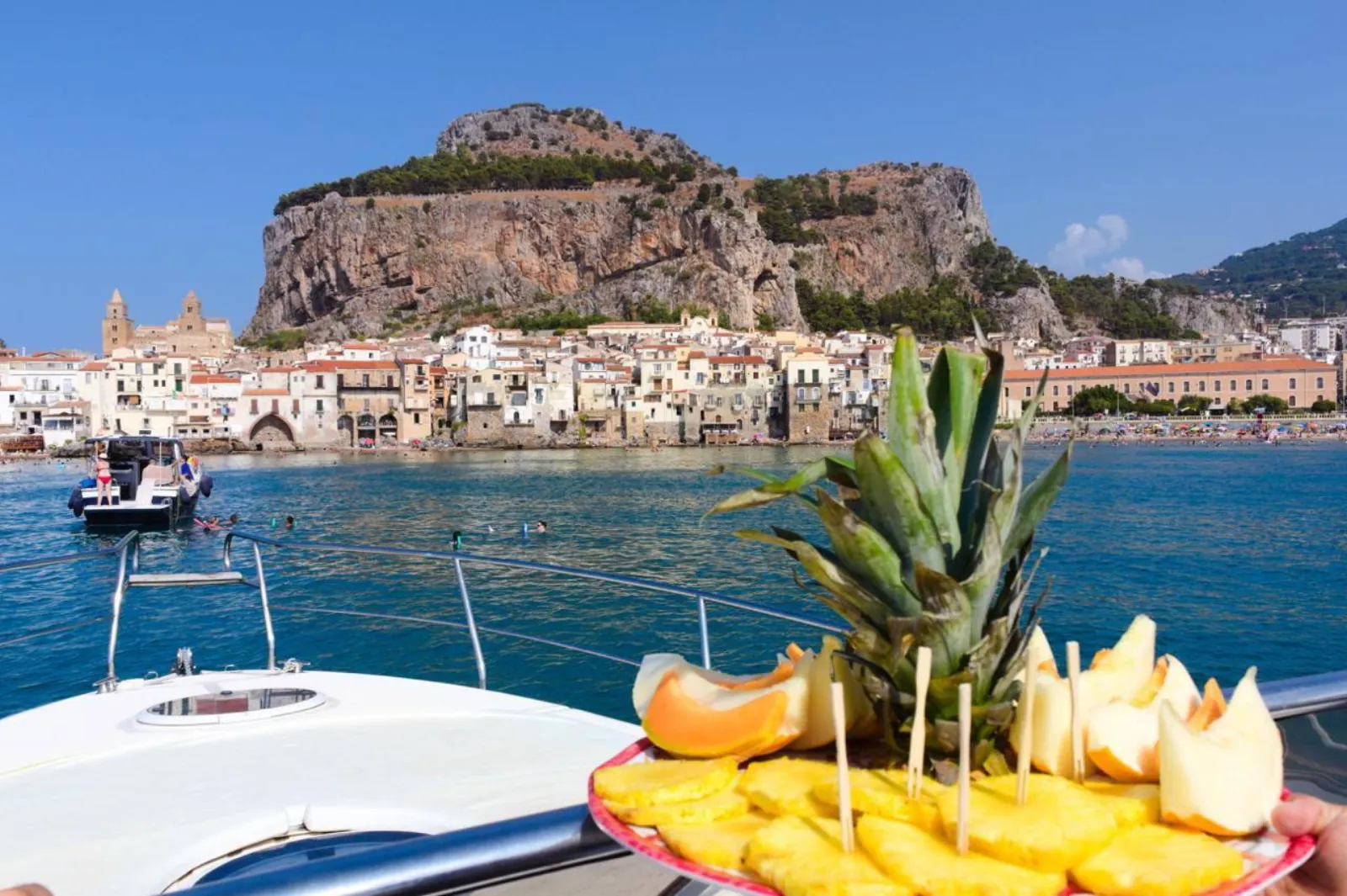 A platter of fresh Sicilian fruit set out on the aft deck, with Cefalù's waterfront behind